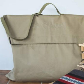 A Glimakra Emilia Rigid Heddle Loom Bag in beige, featuring a rectangular shape, flap closure, and carrying handle, sits on a surface. The bag showcases the stitched Glimakra logo on the front. In the bottom right corner, part of a knitted fabric with striped patterns in red, white, and black is visible—a perfect companion for your Glimakra Emilia rigid heddle loom.