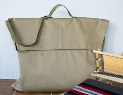 A large, beige Glimakra Emilia Rigid Heddle Loom Bag with a handle and strap is partially open, labeled "Chimayo." Next to the bag is a wooden-frame Glimakra Emilia rigid heddle loom with woven fabric featuring black, white, red, and blue stripes. The scene is set against a plain background.