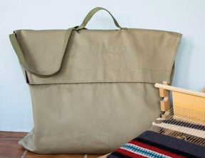 A large, olive green Glimakra Emilia Rigid Heddle Loom Bag with a top flap closure is shown against a light blue background. The bag features a sturdy strap for carrying. To its right, part of a colorful woven blanket created with the Glimakra Emilia rigid heddle loom is partially visible.