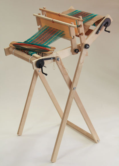 A Glimakra Emilia Rigid Heddle Loom stand made of birch wood, featuring a partially woven fabric. The loom is set up with green and blue threads and showcases solid metal gears along with various wooden components. It stands on three legs, providing a stable base for the loom, which comes complete with weaving instructions.