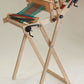 A Glimakra Emilia Rigid Heddle Loom by Glimakra stands on a tripod. The loom features intricate mechanisms with solid metal gears and levers, and it is partially threaded with colorful yarn. The yarn weaves a pattern of green, blue, and red stripes, following detailed weaving instructions.