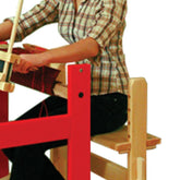 A person with blonde hair is sitting on the Glimakra Julia Loom Bench made from solid wood. They are operating a red loom and weaving fabric, dressed in a plaid shirt and black pants, with an intense focus reminiscent of traditional Swedish craftsmen.