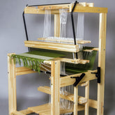 A Glimakra Julia 26" 8-shaft countermarch loom in a natural finish, with green and white threads arranged on it. The loom features multiple beams and levers, with the green threads forming a partially completed woven fabric. The overall setup is clean and organized against a plain gray background.