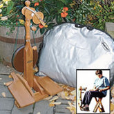 The Majacraft Little Gem Spinning Wheel from Majacraft Co. stands on a patio next to a large, silver carrying case. The surroundings include a wooden barrel and green foliage. An inset shows a person demonstrating how to use the lightweight spinning wheel, sitting and engaged in spinning.