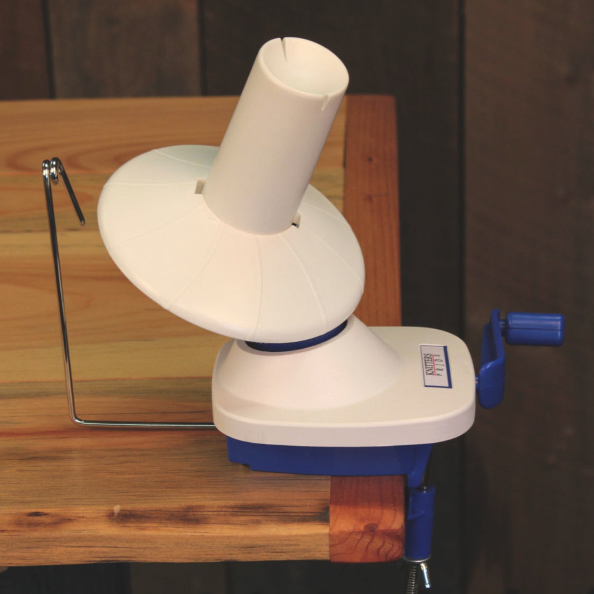 An Accessories Unlimited Knitter's Pride Ball / Wool Winder is clamped to the edge of a wooden table. The device features a white plastic cone, round base, and blue winding handle. A metal wire arm is elevated on the left side to guide the yarn. The background appears to be wooden paneling.