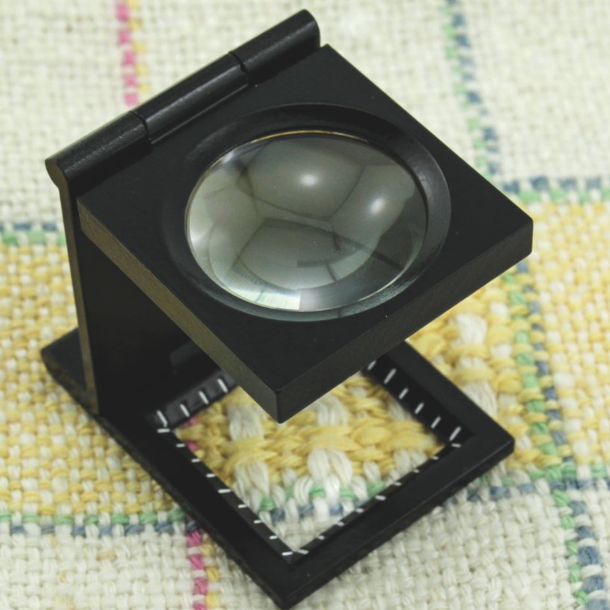 A black folding Magnifying Pick Counter Glass with Rule from Edmunds Optical sits on a textured, woven fabric background with yellow, green, and pink threads. The tool is positioned open, showcasing its lens and rectangular base.