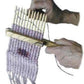 Hands weaving lilac-colored yarn on a Lacis 12 Stick Walking Stick-Loom. The walking stick-loom from Lacis is held in one hand while the other guides the yarn through the threads, effortlessly creating a patterned tapestry weave.