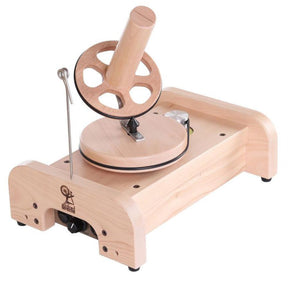 The Ashford Wooden Ball Winder by Ashford Handicrafts Limited features a horizontal wooden base, a rotating wheel with spokes, and a central winding post. A metal guide arm extends vertically from the base to help direct the yarn onto the winder. The winder also displays the brand logo of Ashford Handicrafts Limited.