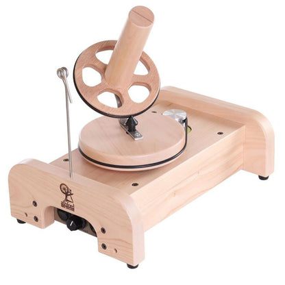 The Ashford Wooden Ball Winder by Ashford Handicrafts Limited features a horizontal wooden base, a rotating wheel with spokes, and a central winding post. A metal guide arm extends vertically from the base to help direct the yarn onto the winder. The winder also displays the brand logo of Ashford Handicrafts Limited.