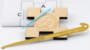 A small square wooden "Weaving Sett Tool" by Halcyon Yarn, featuring the image of a bird and text, along with a blue plastic needle and a gold-colored metal hook with engraved text, are displayed against a white background. Additionally, part of a paper with letters and lines is visible in the background, likely for testing shrinkage.