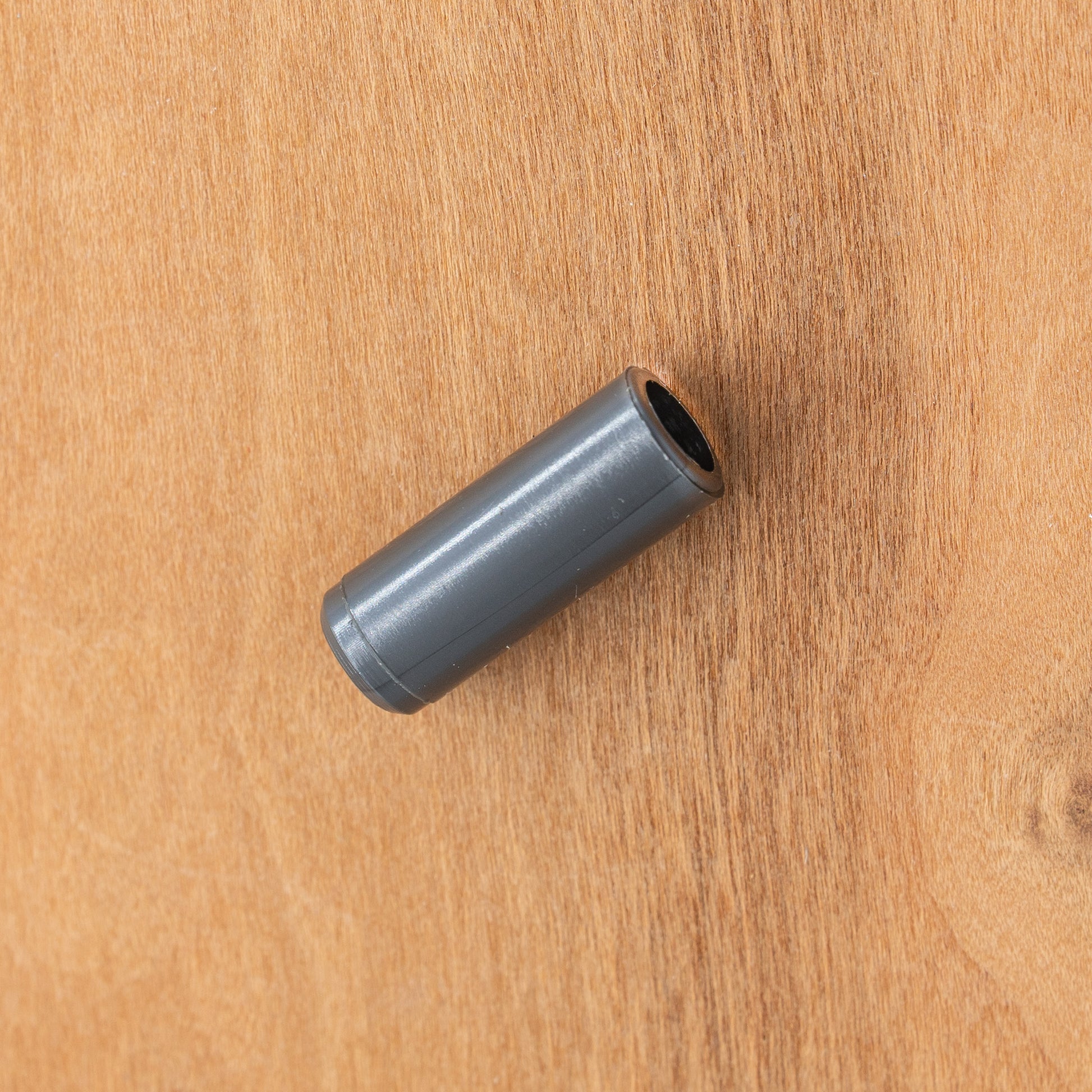 An Ashford Maiden Bearing, a small cylindrical metal object from Ashford Handicrafts Limited, is placed on a wooden surface. The smooth, ashford-gray piece with reflective qualities is positioned slightly angled to the right.