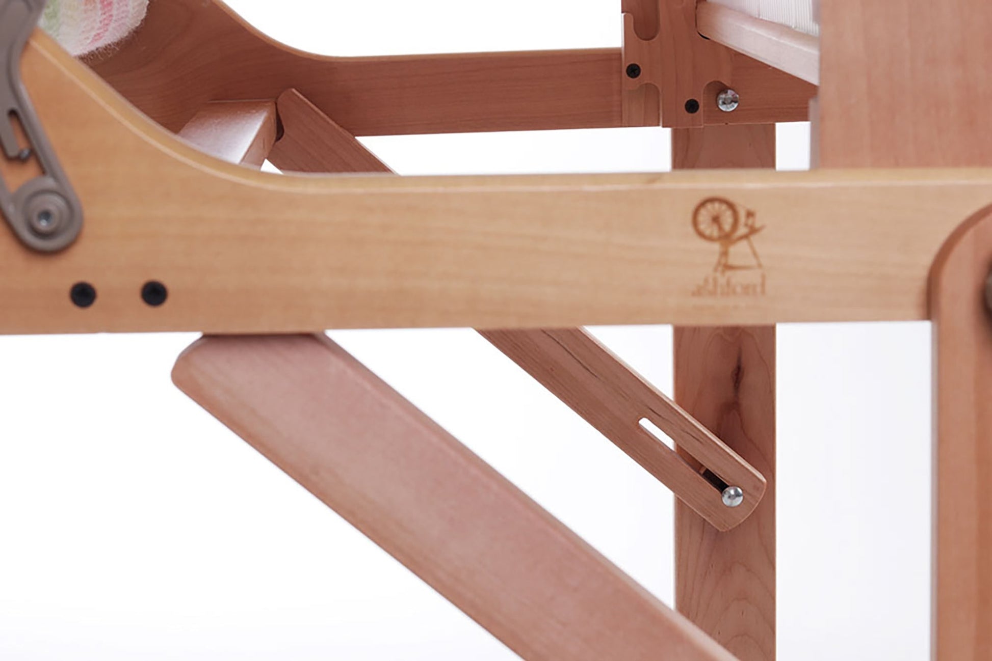 A close-up view of the Ashford Rigid Heddle Table Stand by Ashford Handicrafts Limited reveals a wooden furniture joint with intricate metal hinges. The wood boasts a smooth finish, complemented by an engraved design of a spinning wheel and the word "Stokke" on one of the surfaces. This stand offers an adjustable weaving angle, all set against a plain, soft white background.