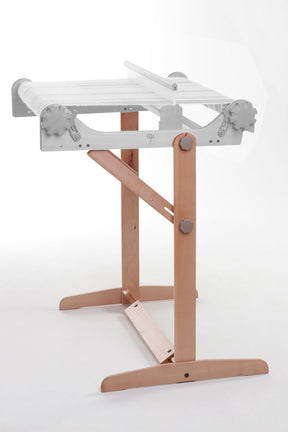 The Ashford Rigid Heddle Table Stand by Ashford Handicrafts Limited is a weaving stand crafted from wood and metal. This stand includes adjustable knobs for controlling height and tension, a sturdy wooden base with two legs for stability, and a metal frame on top with a roller to hold the weaving material. Its minimalistic design also offers an adjustable weaving angle for added convenience.