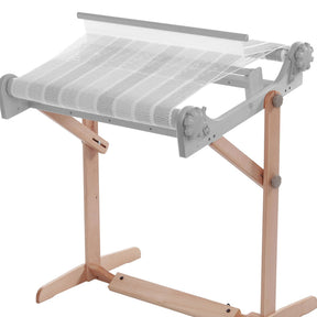 The Ashford Rigid Heddle Table Stand by Ashford Handicrafts Limited is a weaving loom with a wooden stand and a metal frame. It features white threads partially woven into fabric, positioned at an adjustable weaving angle. The wooden base ensures stability for the loom.