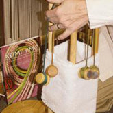 A person is demonstrating or assembling a Lacis Kumihimo Stand with a white fabric attached and colored threads hanging from it. In the background, a colorful pattern book or magazine titled "Kumihimo" complements the scene, resembling how one might use the stand for intricate patterns.