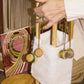 A person is demonstrating or assembling a Lacis Kumihimo Stand with a white fabric attached and colored threads hanging from it. In the background, a colorful pattern book or magazine titled "Kumihimo" complements the scene, resembling how one might use the stand for intricate patterns.