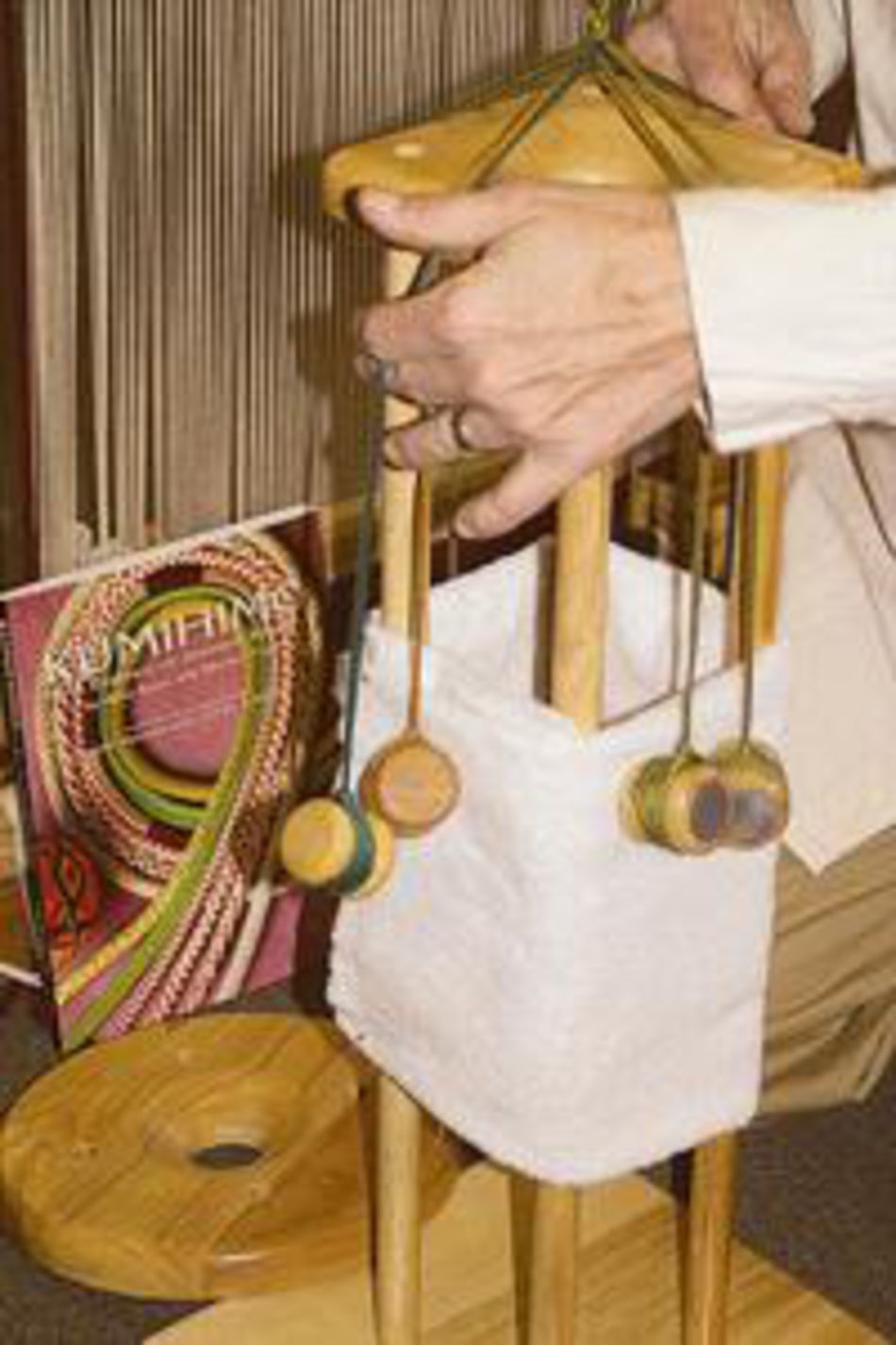 A person is demonstrating or assembling a Lacis Kumihimo Stand with a white fabric attached and colored threads hanging from it. In the background, a colorful pattern book or magazine titled "Kumihimo" complements the scene, resembling how one might use the stand for intricate patterns.