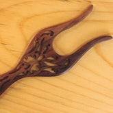 A handcrafted Filigree Rosewood Lucet by Lacis, featuring intricate carved designs, lies on a light wooden surface. This hair fork, made from rosewood lucet, boasts two elongated and curved prongs that showcase a finely detailed ornamental pattern in the center.