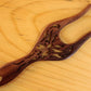 A dark brown, intricately carved Filigree Rosewood Lucet by Lacis with a hole at one end is placed on a light wood background. The finely detailed hair fork features decorative cutouts and has a polished finish.