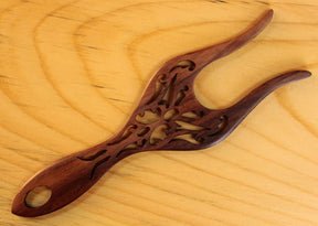 A dark brown, intricately carved Filigree Rosewood Lucet by Lacis with a hole at one end is placed on a light wood background. The finely detailed hair fork features decorative cutouts and has a polished finish.