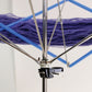 Close-up of the Lacis Plastic and Metal Swift/Skein Winder with an adjustable clamp, holding a skein of purple yarn. The swift's extendable arms crisscross to keep the yarn secure, ready for winding skeins. The background is a neutral, out-of-focus wall.