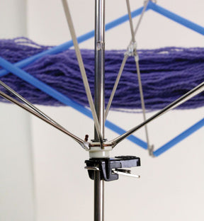 Close-up of the Lacis Plastic and Metal Swift/Skein Winder with an adjustable clamp, holding a skein of purple yarn. The swift's extendable arms crisscross to keep the yarn secure, ready for winding skeins. The background is a neutral, out-of-focus wall.