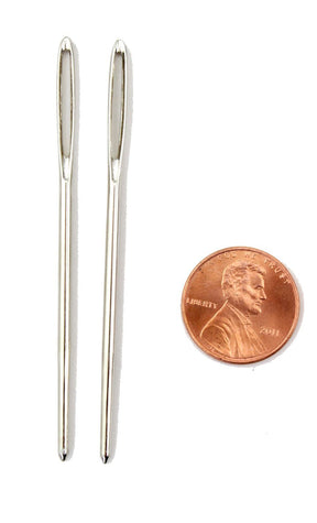 Two Large-Eyed Tapestry Needles from Accessories Unlimited are placed vertically next to a U.S. penny, which features the profile of Abraham Lincoln. The metallic needles, slightly larger than the penny, stand out against the white background, highlighting their shiny appearance and size ideal for bulky weight yarns.