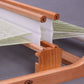 A close-up view of a small wooden weaving loom in use reveals intricate details. The Ashford Second Rigid Heddle Support Block Kit by Ashford Handicrafts Limited features white and green threads stretched across its frame, with a comb-like structure and gears prominently displayed, demonstrating the weaving process against a plain gray background.