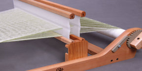 A close-up view of a small wooden weaving loom in use reveals intricate details. The Ashford Second Rigid Heddle Support Block Kit by Ashford Handicrafts Limited features white and green threads stretched across its frame, with a comb-like structure and gears prominently displayed, demonstrating the weaving process against a plain gray background.