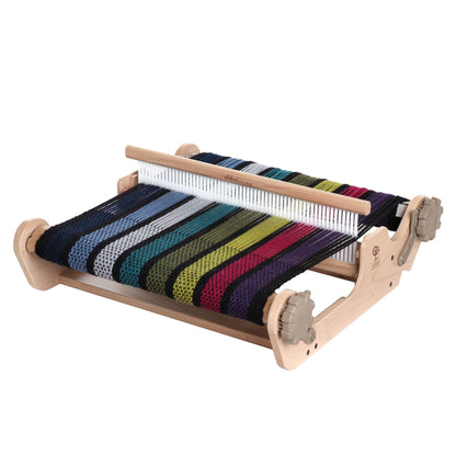 An Ashford SampleIt Rigid Heddle Loom with a partially woven fabric showcasing vibrant stripes in black, blue, green, yellow, and pink. This weaving tool from Ashford Handicrafts Limited features metal heddles, tension knobs, and a sturdy rectangular frame – essential supplies for any craft enthusiast.