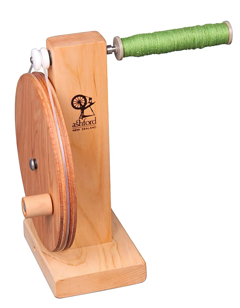 An Ashford Boat Shuttle Bobbin Winder Wooden with a natural wooden finish from Ashford Handicrafts Limited. The device features a large wooden wheel on one side and a stainless steel shaft with nylon bearings supporting the spool of green yarn on the other. Its sturdy base provides stability during use, and the logo reads "Ashford New Zealand.