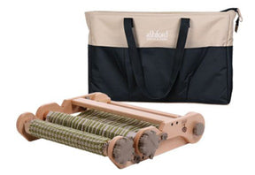 An Ashford Rigid Heddle Knitters Loom with partially woven green and white fabric. This loom, crafted by Ashford Handicrafts Limited, is made of light-colored wood and includes reels for the yarn. Behind the loom is a two-tone beige and black canvas carrying bag, complete with handles and a pocket. The package also includes a second heddle kit.