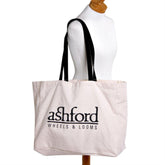 An Ashford Cotton Canvas Carry Bag for SampleIt Looms with black handles hangs on a white mannequin. This attractive, off-white bag features the text "ashford WHEELS & LOOMS" printed in black letters. The background is plain white.