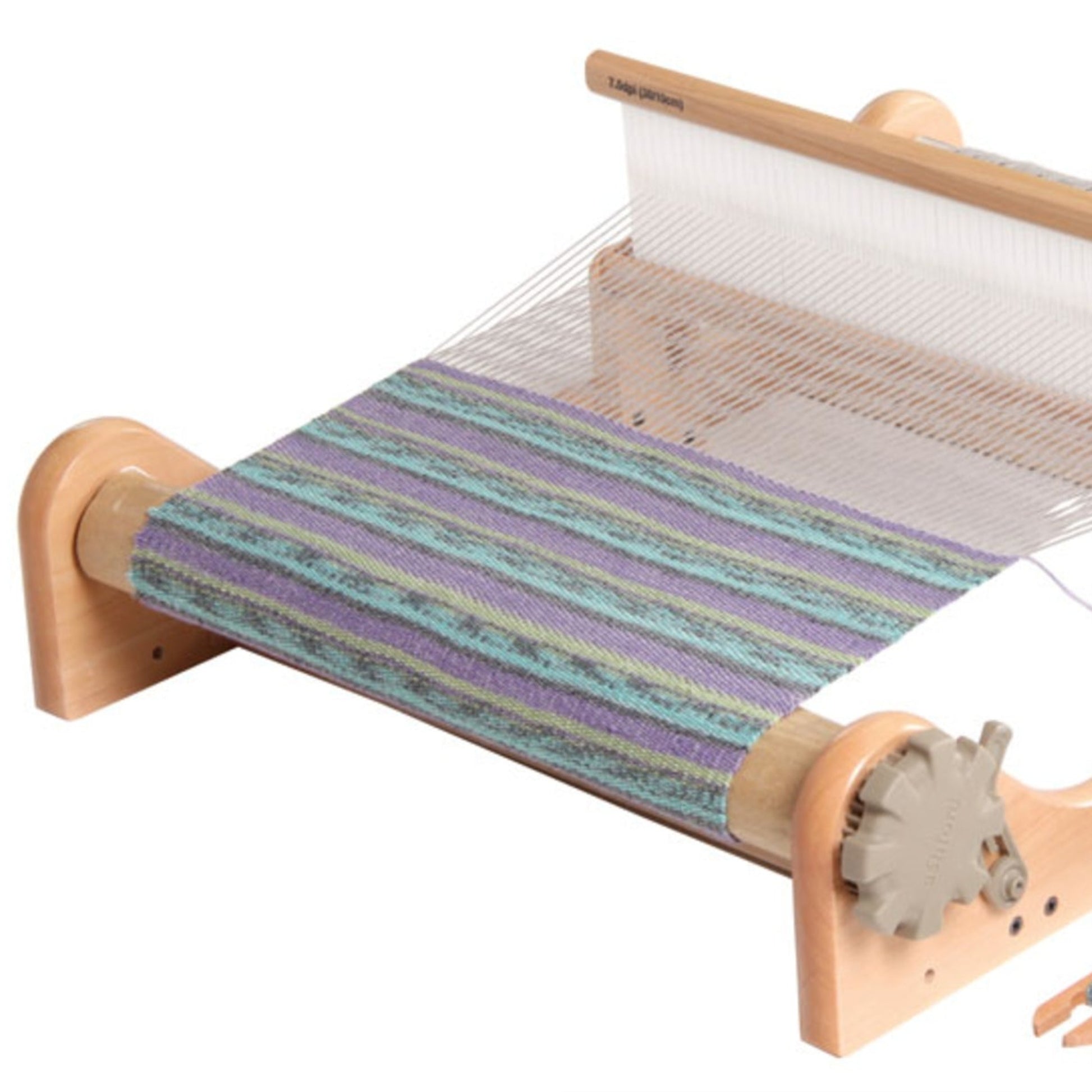 The Ashford Rigid Heddle Loom by Ashford Handicrafts Limited is a small wooden table loom with a partially woven fabric featuring blue, green, and purple stripes. This rigid heddle loom boasts a light wood frame and a metal gear on the side for adjusting tension, beautifully showcasing the art of hand weaving in a portable format.