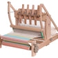 An Ashford Table Loom with an Overhead/Swinging Beater from Ashford Handicrafts Limited intricately weaves colorful horizontal striped fabric, featuring blue, green, yellow, and orange hues. Various mechanical components such as gears and Texsolv heddles are visible to manage the weaving process.