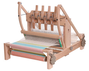 An Ashford Table Loom with an Overhead/Swinging Beater from Ashford Handicrafts Limited intricately weaves colorful horizontal striped fabric, featuring blue, green, yellow, and orange hues. Various mechanical components such as gears and Texsolv heddles are visible to manage the weaving process.