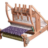 An Ashford Handicrafts Limited Table Loom, featuring an overhead/swinging beater and Texsolv heddles. It is set up with white warp threads and showcases multiple levers for easy operation. The loom has a partially completed purple and white woven fabric on the upper section and a green and white woven fabric rolled up on the lower section.