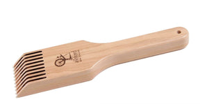 The Ashford Tapestry Beater, crafted from Silver Beech hardwood and featuring a lacquer finish, includes a handle adorned with a carved bicycle and the brand name "Ashford Handicrafts Limited." This loom accessory is designed with multiple closely spaced teeth and has a small hole at the end of the handle, which may be used for hanging.