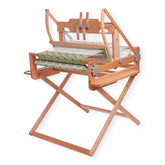 The Ashford Stand for Table Loom by Ashford Handicrafts Limited features a robust wooden frame with green and white patterned fabric partially woven on it. This loom is equipped with levers and gears, mounted on a foldable stand with two shelves for convenient storage.