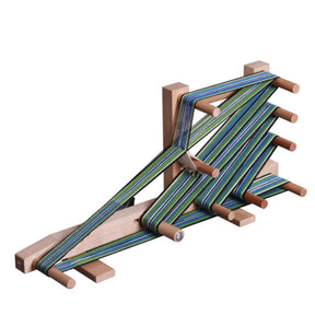 A wooden weaving tool, specifically a warp board, features multiple pegs and green-blue striped yarn meticulously wrapped around it in an intricate pattern. Often used in conjunction with the Ashford Inkle Loom by Ashford Handicrafts Limited, this board is designed to measure and prepare yarn for weaving projects like warp-faced straps or belts.