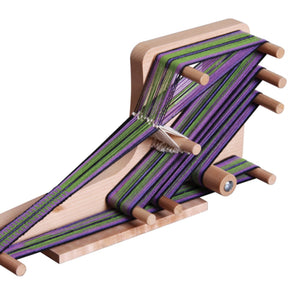 A lightweight Ashford Inklette Loom by Ashford Handicrafts Limited, features a weaving project in progress. The loom holds an intricate pattern of green and purple threads interwoven tightly around several wooden pegs. This portable setup is compact and rests on a smooth wooden base, ready to accommodate various weaving lengths.