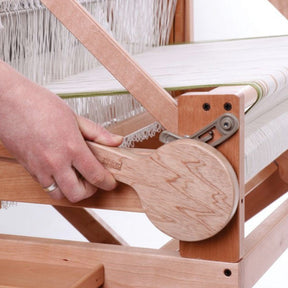 Close-up of a person’s hand adjusting an Ashford Handi Handle on an Ashford loom. Strings of thread are stretched across the loom, ready for weaving. The craftsman’s hand firmly grips the tool to facilitate the weaving process, demonstrating careful control even for those with impaired hand mobility.