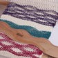 A close-up of a weaving project on a loom showcases decorative patterns with alternating bands of purple, white, teal, and red threads. A wooden weaving comb branded "Schacht" and an Ashford Wavy Stick Shuttle by Ashford Handicrafts Limited rest across the woven fabric, highlighting the intricate textured designs.