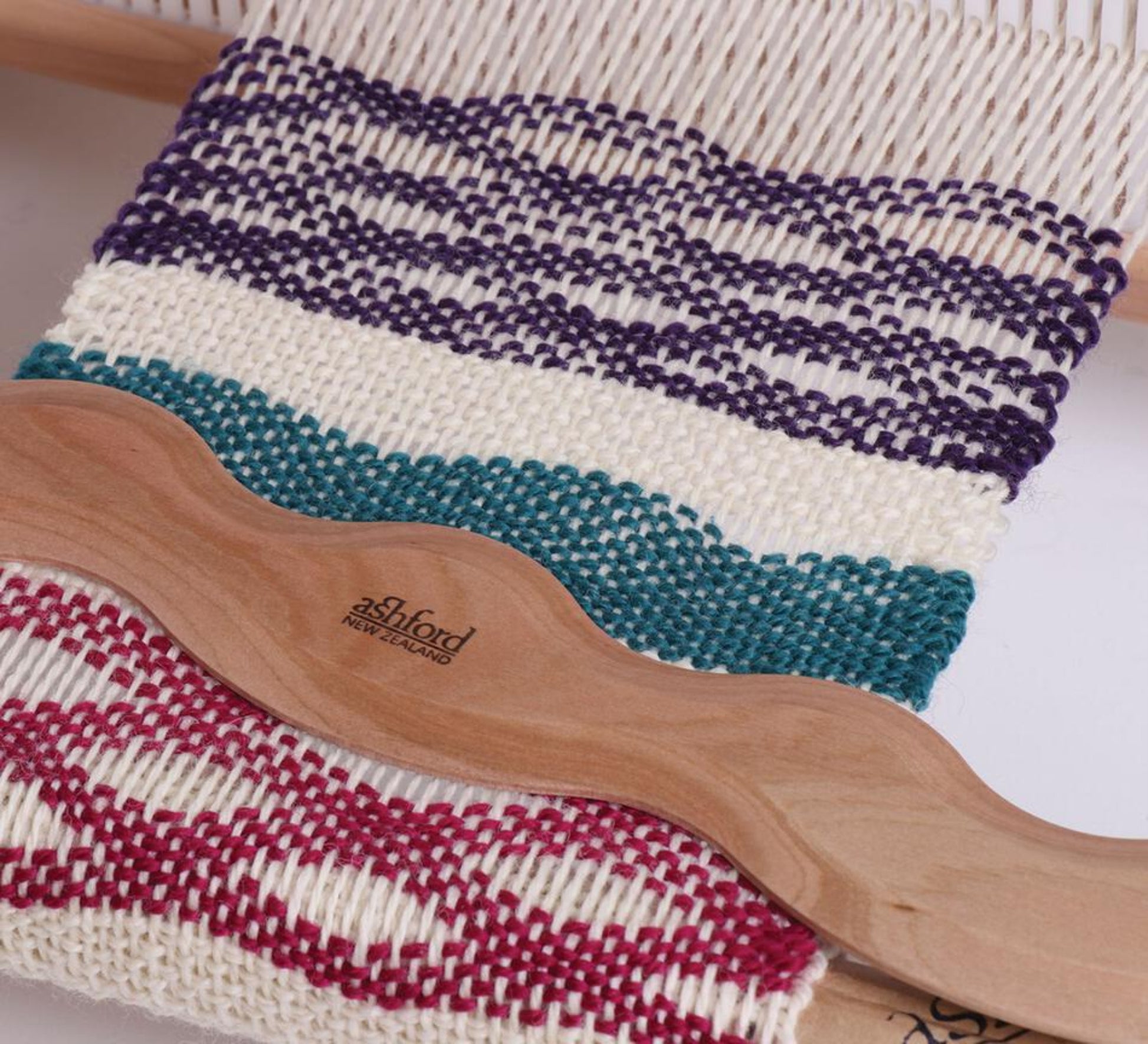 A close-up of a weaving project on a loom showcases decorative patterns with alternating bands of purple, white, teal, and red threads. A wooden weaving comb branded "Schacht" and an Ashford Wavy Stick Shuttle by Ashford Handicrafts Limited rest across the woven fabric, highlighting the intricate textured designs.