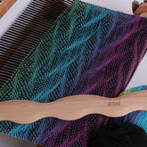 A wooden weaving loom with a vibrant, multicolored woven cloth featuring a gradient of green, blue, purple, and magenta hues in a chevron pattern. Resting on the loom is an Ashford Wavy Stick Shuttle from Ashford Handicrafts Limited—an essential accessory that enhances the decorative patterns in the fabric.