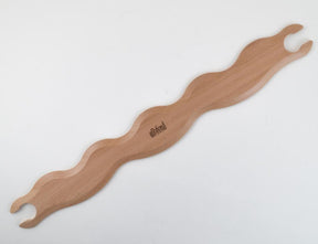 A wooden massage tool with a wavy design, featuring two semi-circular notches at each end. The Ashford Wavy Stick Shuttle from Ashford Handicrafts Limited boasts a smooth finish and decorative patterns, along with the brand’s logo engraved in the center. The tool is displayed on a light-colored surface.