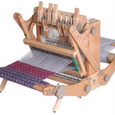 The Ashford Katie, 8-Shaft Table Loom, 12" by Ashford Handicrafts Limited is a wooden tabletop weaving loom with intricate mechanisms and handles. It features partially completed fabric in purple and white patterns. This portable loom has various levers and gears, exemplifying the craft of weaving using its 8-shaft design.