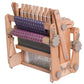 An Ashford Katie, 8-Shaft Table Loom, 12" by Ashford Handicrafts Limited is shown with a partially woven piece of fabric featuring a purple and white geometric pattern. This portable weaving loom has several adjustable knobs and multiple heddles for threads, making it versatile and perfect for intricate designs.