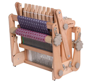 An Ashford Katie, 8-Shaft Table Loom, 12" by Ashford Handicrafts Limited is shown with a partially woven piece of fabric featuring a purple and white geometric pattern. This portable weaving loom has several adjustable knobs and multiple heddles for threads, making it versatile and perfect for intricate designs.