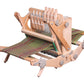 The Ashford Katie, an 8-shaft table loom from Ashford Handicrafts Limited, features a portable wooden frame with green and brown yarn threads. This loom includes several levers, knobs for adjustments, and multiple heddles. The partially completed fabric extends from the front.